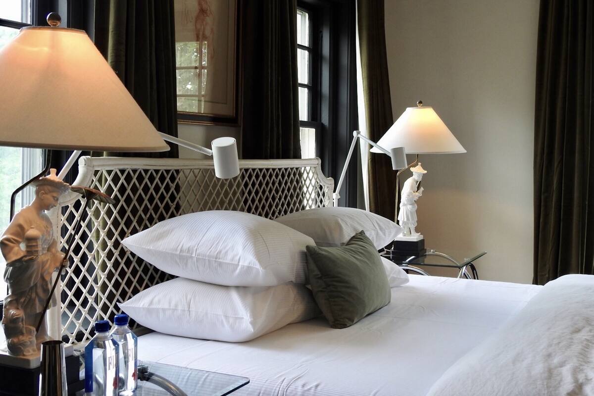 Habitación de hotel con una cama bien hecha, dos lámparas de noche, una estatua decorativa y un ventanal con cortinas.