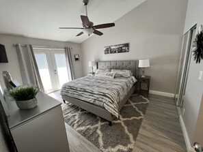 A bedroom with a bed, a ceiling fan, a nightstand, a lamp, a potted plant, and a view of the outdoors.