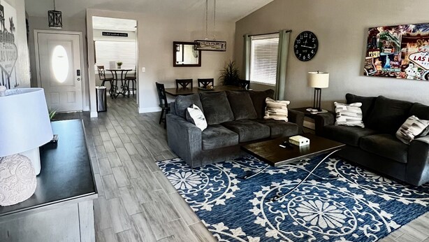 Un salon spacieux avec un canapé de couleur gris foncé, une table basse et un tapis à motifs. On aperçoit à l’arrière-plan un coin repas avec une table et des chaises.
