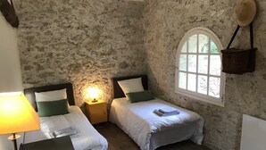 A rustic room with two beds, a nightstand with a lamp, a window with a view, and a woven basket on the wall. - Demeure de Charme 10 Personnes Avec Piscine (Sainte-Colombe-en-Bruilhois)