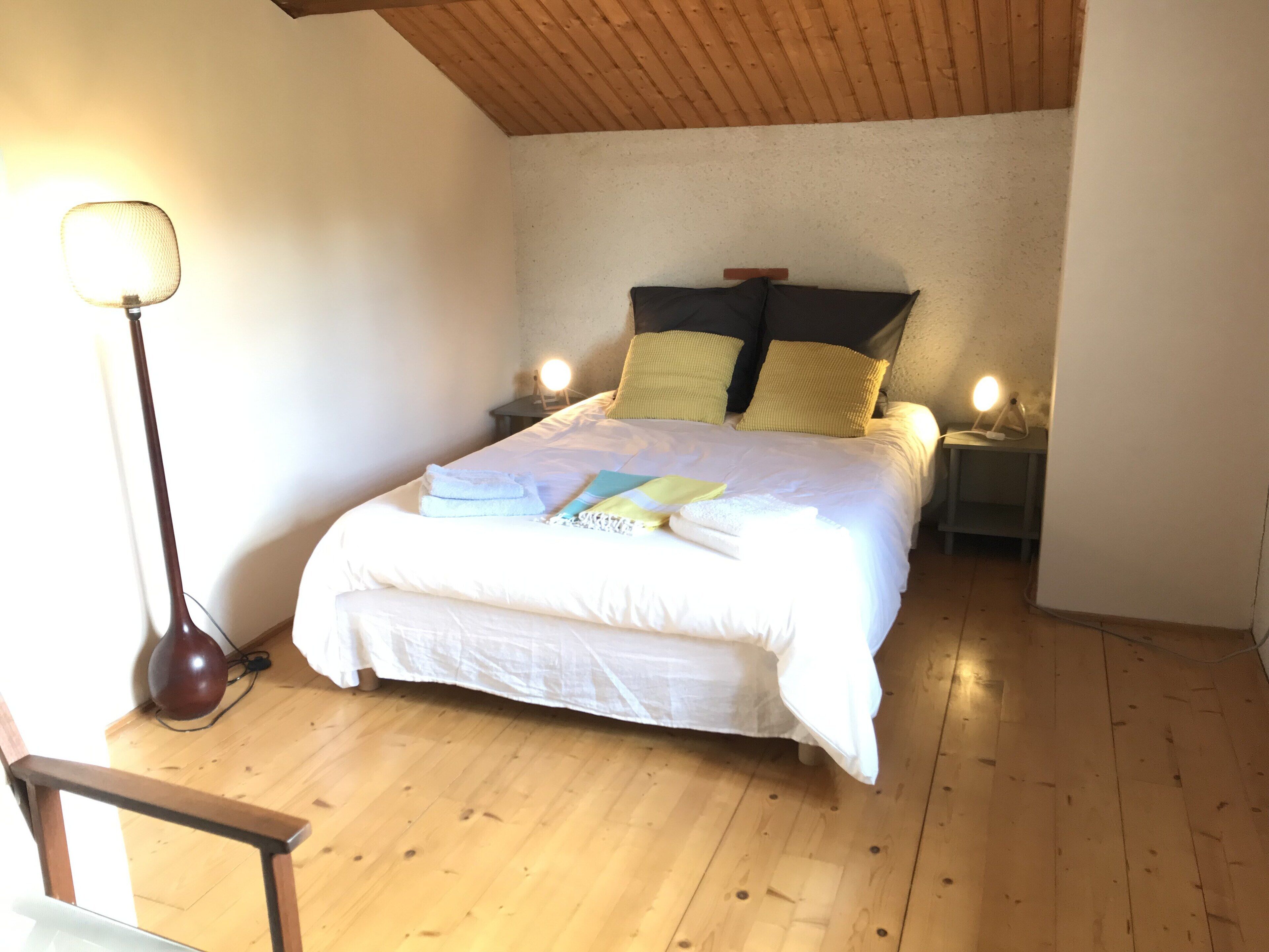 Un dormitorio con suelo de madera, una cama con ropa de cama blanca, dos lámparas de noche y una lámpara de pie.