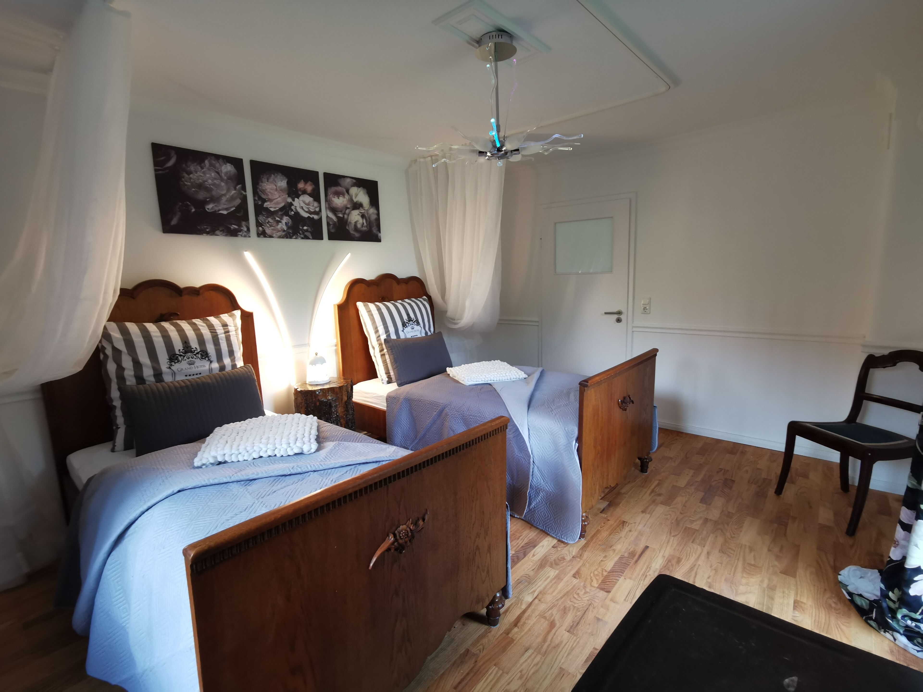 Un dormitorio con dos camas de madera, cortinas blancas, un ventilador de techo y una silla de madera.