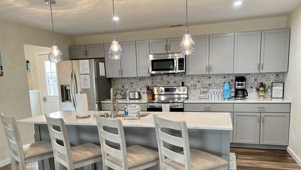 A modern kitchen with grey cabinets, a central island with bar stools, stainless steel appliances, and pendant lighting. - Perfect Location for your Beach/Lake MI stay!  You'll Love This Beautiful Home! (New Buffalo)