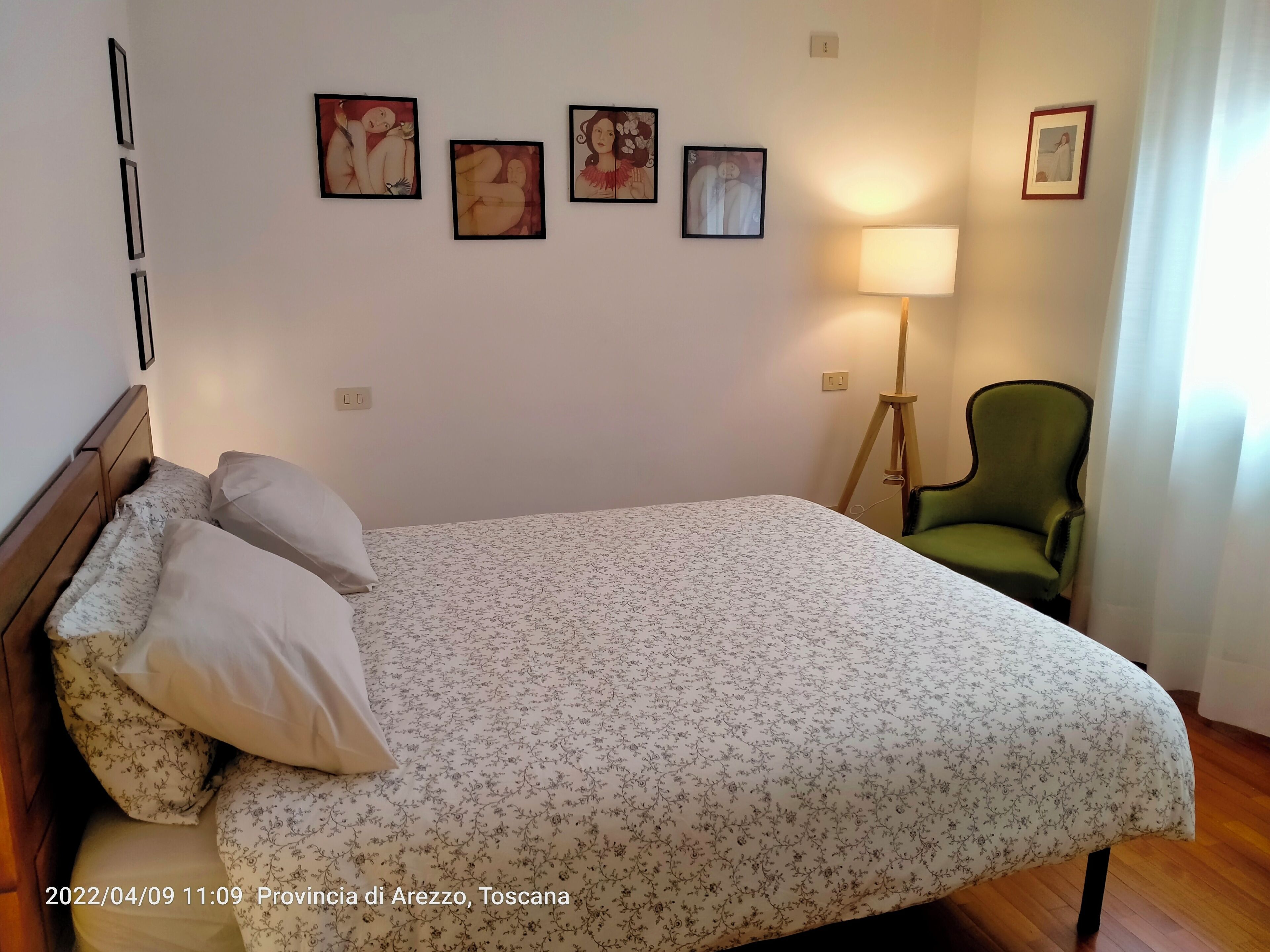 Cama bem arrumada com roupa de cama floral, uma poltrona verde e arte nas paredes em um quarto com piso de madeira.