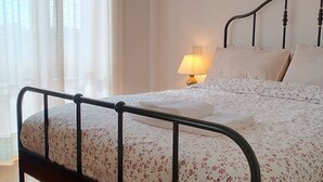 Una cama con cubrecama estampado con motivos florales, una lámpara de noche y una ventana con cortinas de gasa.