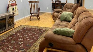 Ein Wohnzimmer mit braunem Sofa, Holzboden, Teppich, einem Holztisch, einem Fernseher und einer Küchenzeile mit Schränken und Geräten.