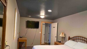 Ein Schlafzimmer mit einem Bett, einem Schreibtisch, einem an der Wand montierten Fernseher und einer Tür zu einem anderen Raum.