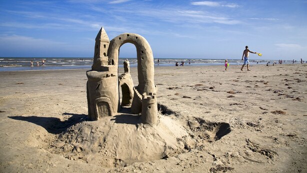 Una playa arenosa con una gran estructura de castillo de arena, que incluye una torre puntiaguda y una entrada arqueada.