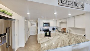 Una cocina moderna con armarios blancos, una isla de mármol, electrodomésticos de acero inoxidable y un letrero que dice 'BEACH HOUSE'.