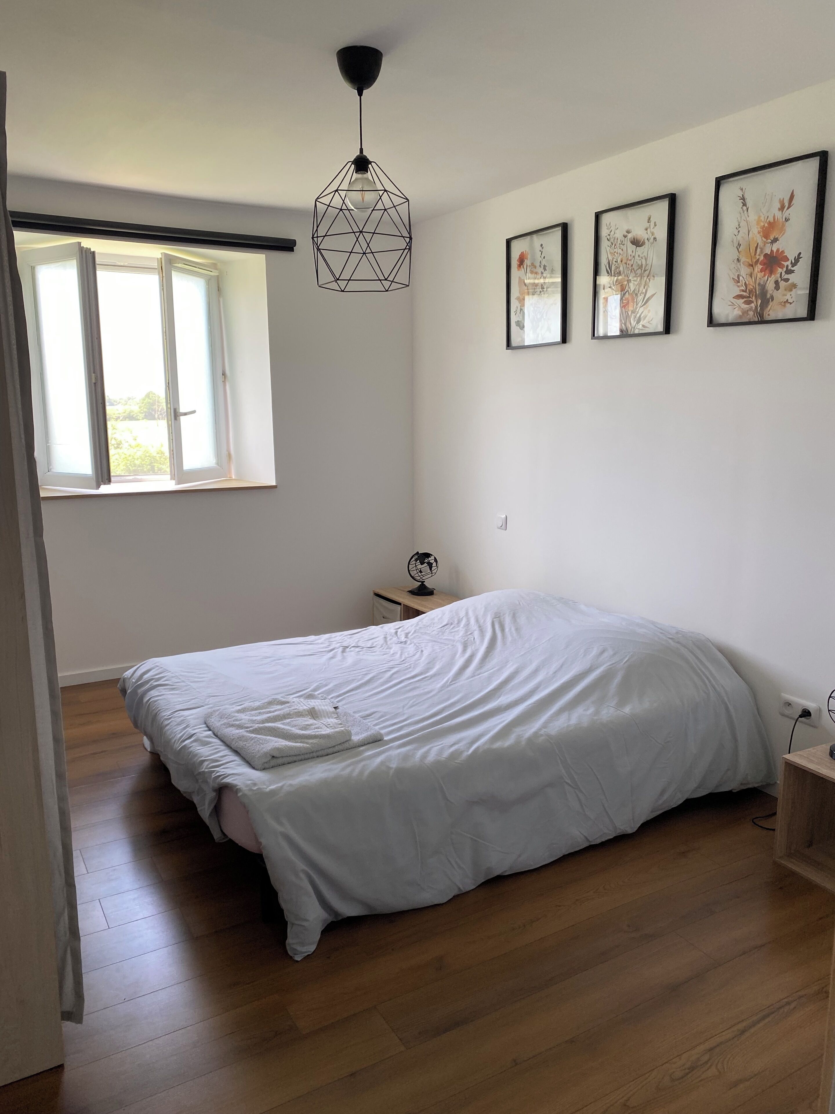 Un dormitorio con una cama grande, una lámpara colgante, un ventanal y tres cuadros con motivos florales en la pared.