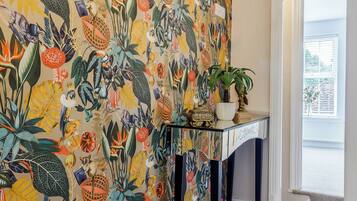 Una habitación con un llamativo papel tapiz floral, una pequeña consola con una planta en maceta y una ventana con persianas.