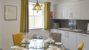A dining area with a round glass table set for six, surrounded by chairs, in a modern kitchen with white cabinets and a window with curtains.