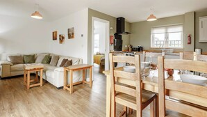 A spacious living area with a sofa, wooden chairs, a coffee table, and pendant lighting. A kitchen with a stove and sink is visible in the background.