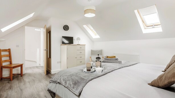 A modern bedroom with a sloped ceiling, a bed with a grey quilt, a wooden chair, a bedside table with a lamp, a TV, and a clock on the wall.