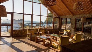 A room with large windows overlooking a beach, wooden furniture, and woven ceiling lamps. - Lukuba Island Lodge (Musoma)