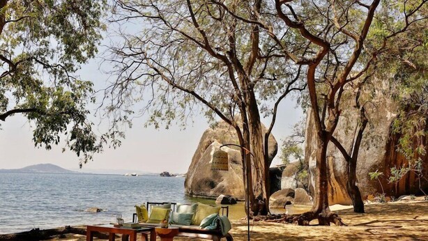 A beach with a wooden table and chairs, a hammock, and a large rock formation in the background. - Lukuba Island Lodge (Musoma)