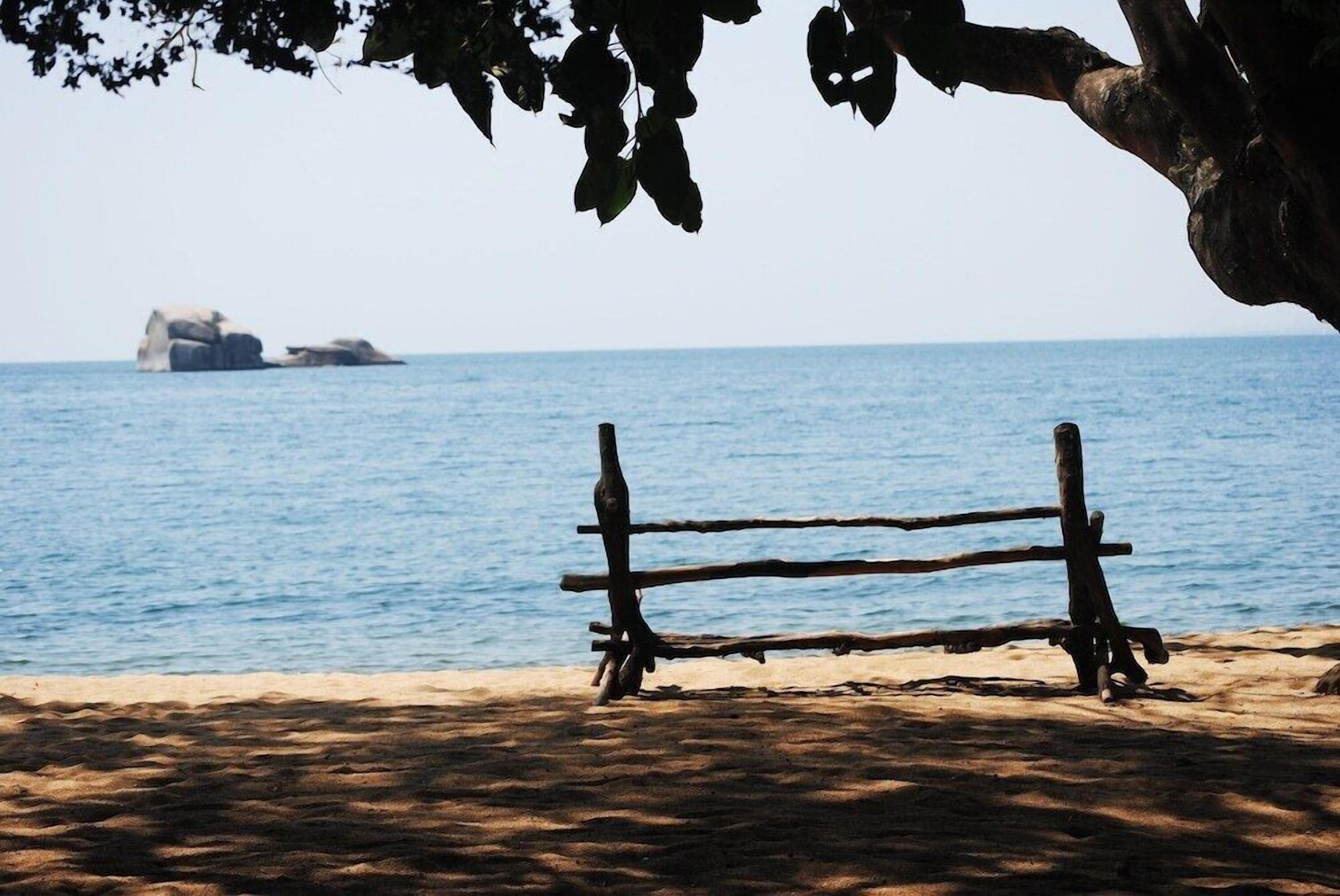 A wooden bench on a sandy beach with a view of the sea and a rock formation in the distance.