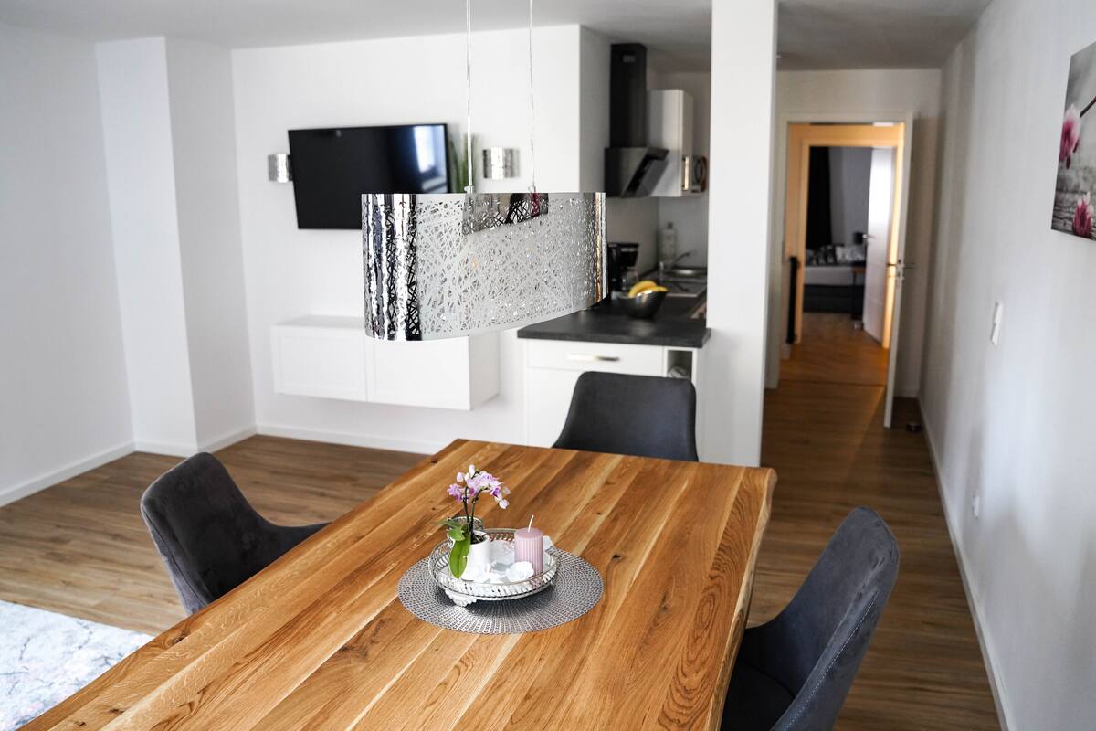 Uma zona de refeições moderna com mesa de madeira, cadeiras pretas e um centro de mesa decorativo.