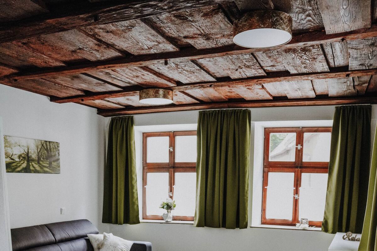 Sala de estar com um sofá escuro, uma mesa pequena branca e um tapete verde. Há duas janelas com cortinas verdes e teto de madeira.