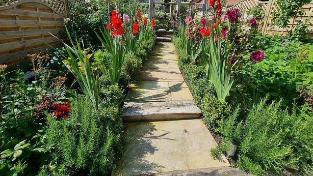 Um caminho no jardim leva a um gazebo com telhado de pedra, rodeado por diversas plantas floridas e vegetação.