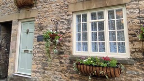 Um edifício de pedra com uma porta verde clara, uma janela grande com cortinas florais e cestos de flores pendurados.