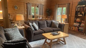A cozy cabin interior with a brown sofa, a matching armchair, a wooden coffee table, and a bookshelf filled with board games. - Beautiful Log Chalet/Cabin with Hot Tub (Berkeley Springs)