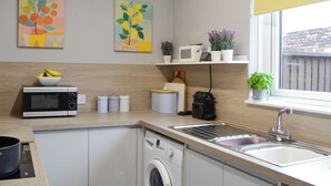 A modern kitchen with a washing machine, microwave, and sink. There are two potted plants on a shelf and a bowl of fruit on the counter. - 1 bedroom accommodation in Tayport, near St Andrews (Tayport, near St Andrews)