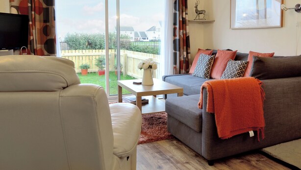 A living room with a sofa, a white armchair, a coffee table, and a TV. There is a sliding glass door leading to a garden. - 1 bedroom accommodation in Tayport, near St Andrews (Tayport, near St Andrews)