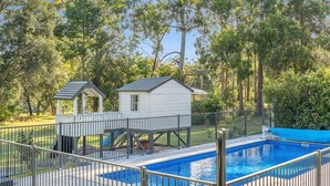 Una piscina con un deck cercado, un pequeño edificio blanco y un área de juegos rodeada de árboles.