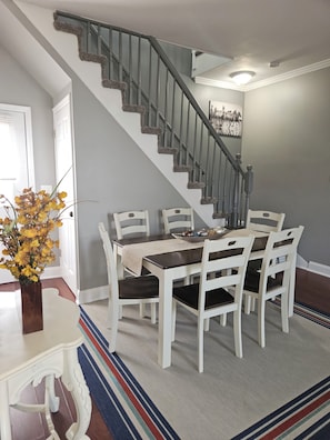 Un comedor con una escalera, sillas blancas, una mesa con mantel beige y un jarrón con flores amarillas.