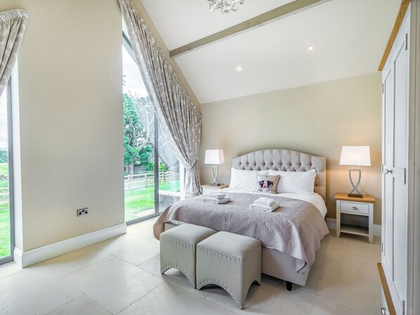 A bedroom with a large bed, two bedside tables with lamps, a bench, and a view of a garden through a window.