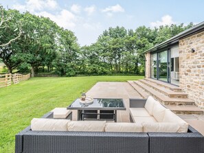A patio with a wicker sofa, a table with a coffee pot, and a stone building with large windows.