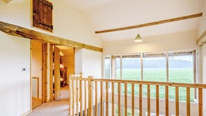 A spacious corridor with a wooden railing, large windows offering a view of a field, and a loft area with exposed beams.