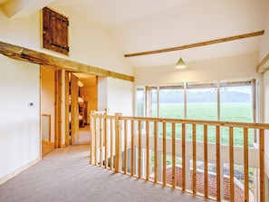 A spacious corridor with a wooden railing, large windows offering a view of a field, and a loft area with exposed beams.