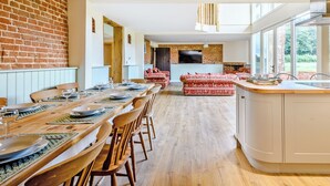 A modern kitchen with a large island, wooden dining table, and chairs. There is a brick wall and a large window providing natural light.