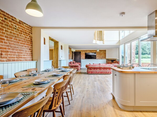 A modern kitchen with a large island, wooden dining table, and chairs. There is a brick wall and a large window providing natural light.