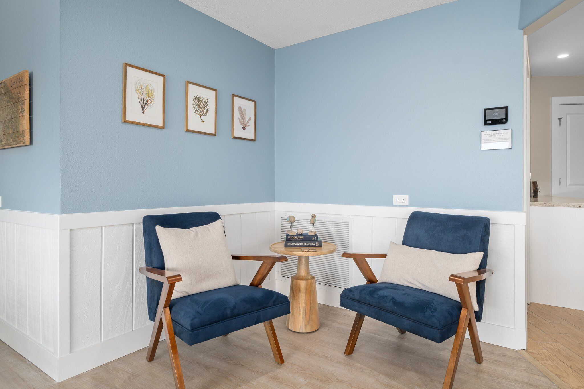 A room with two blue armchairs, a small wooden table, and three framed botanical prints on the wall.