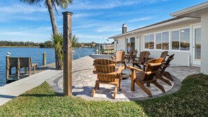 Une terrasse au bord de l’eau, avec des chaises et une table en bois, un palmier, et une vue sur l’eau et les bâtiments situés au loin.