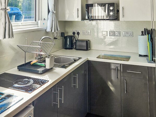Una cocina moderna equipada con microondas, tostadora y hervidor. Hay un lavamanos con un escurreplatos y una ventana con cortinas.