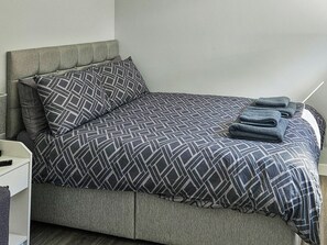 A neatly made bed with a geometric patterned bedspread and pillows, a bedside table with a lamp, and a folded towel on the bed.