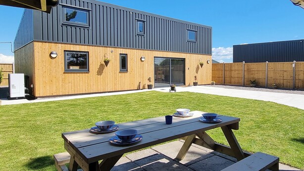 A modern house with a wooden exterior and a grey roof, a patio with a table and chairs, and a well-maintained lawn. - 5 bedroom accommodation in Somerton (Somerton)