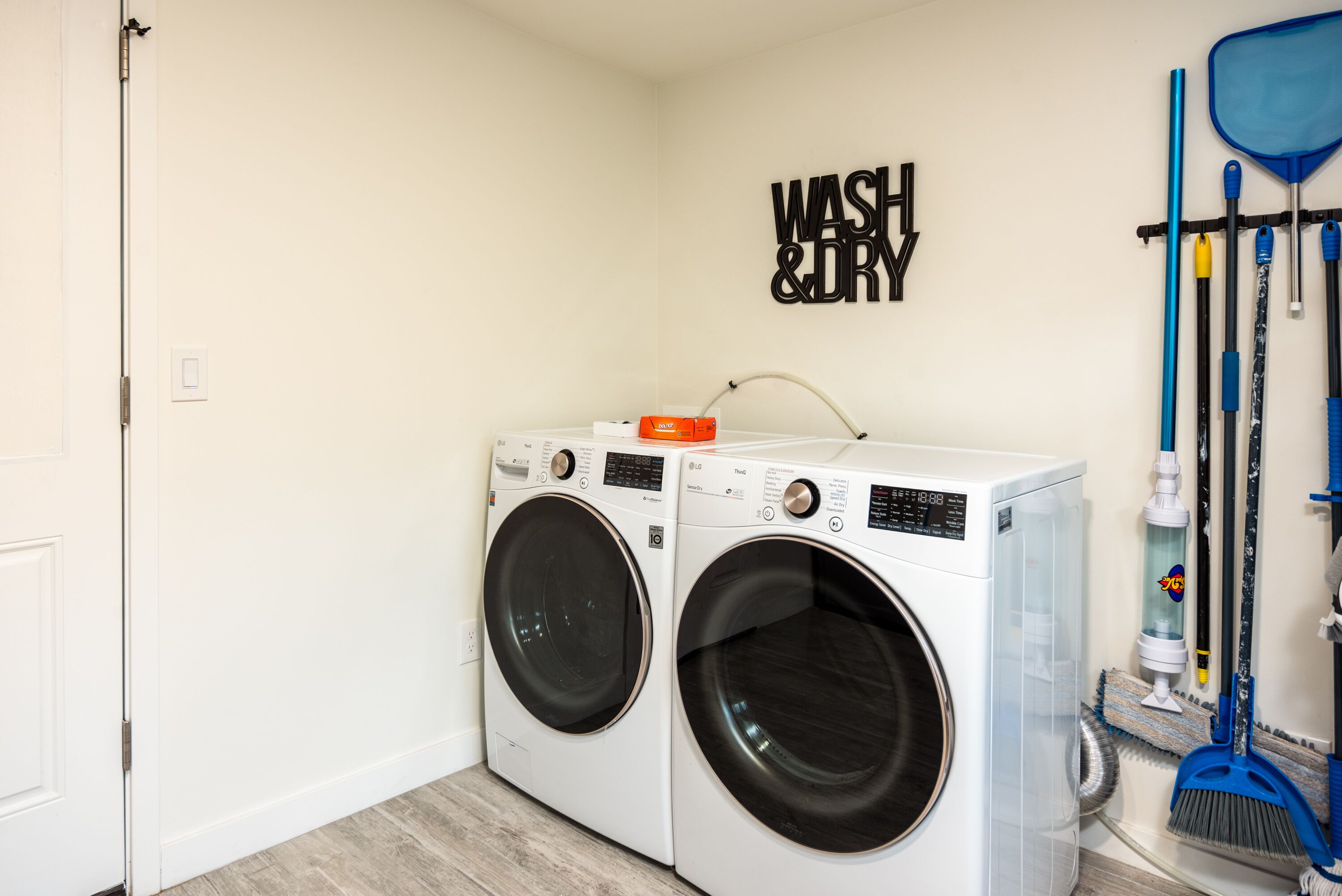 A laundry room with two front-loading washing machines, a wall-mounted "WASH & DRY" sign, and a broom rack with brooms and mops.