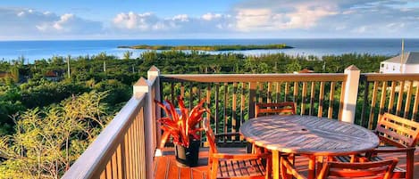 Une terrasse en bois avec une table ronde et des chaises, offrant une vue sur un paysage côtier avec la mer et des îles au loin.