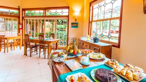 A dining area with a wooden table set for a meal, surrounded by chairs, and large windows with a view of greenery outside. - VELINN Pousada Villa Caiçara (Ilhabela)