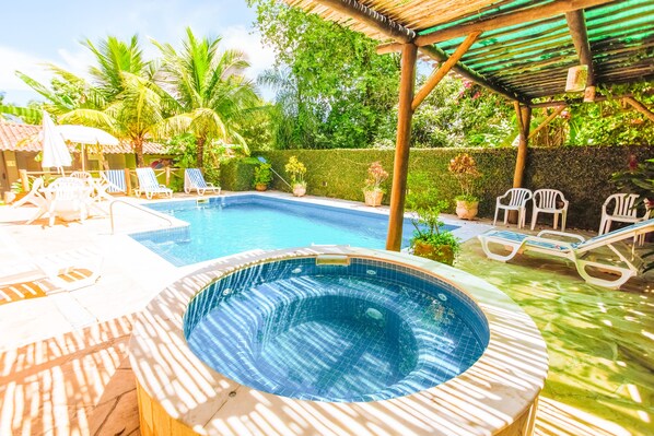 A swimming pool with lounge chairs and a hot tub, surrounded by greenery and a pergola.