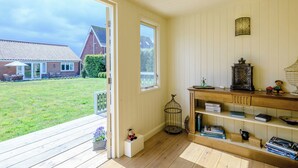 Habitación con piso de madera, una estantería, una mesita y vista a un jardín y una casa a través de una puerta abierta.