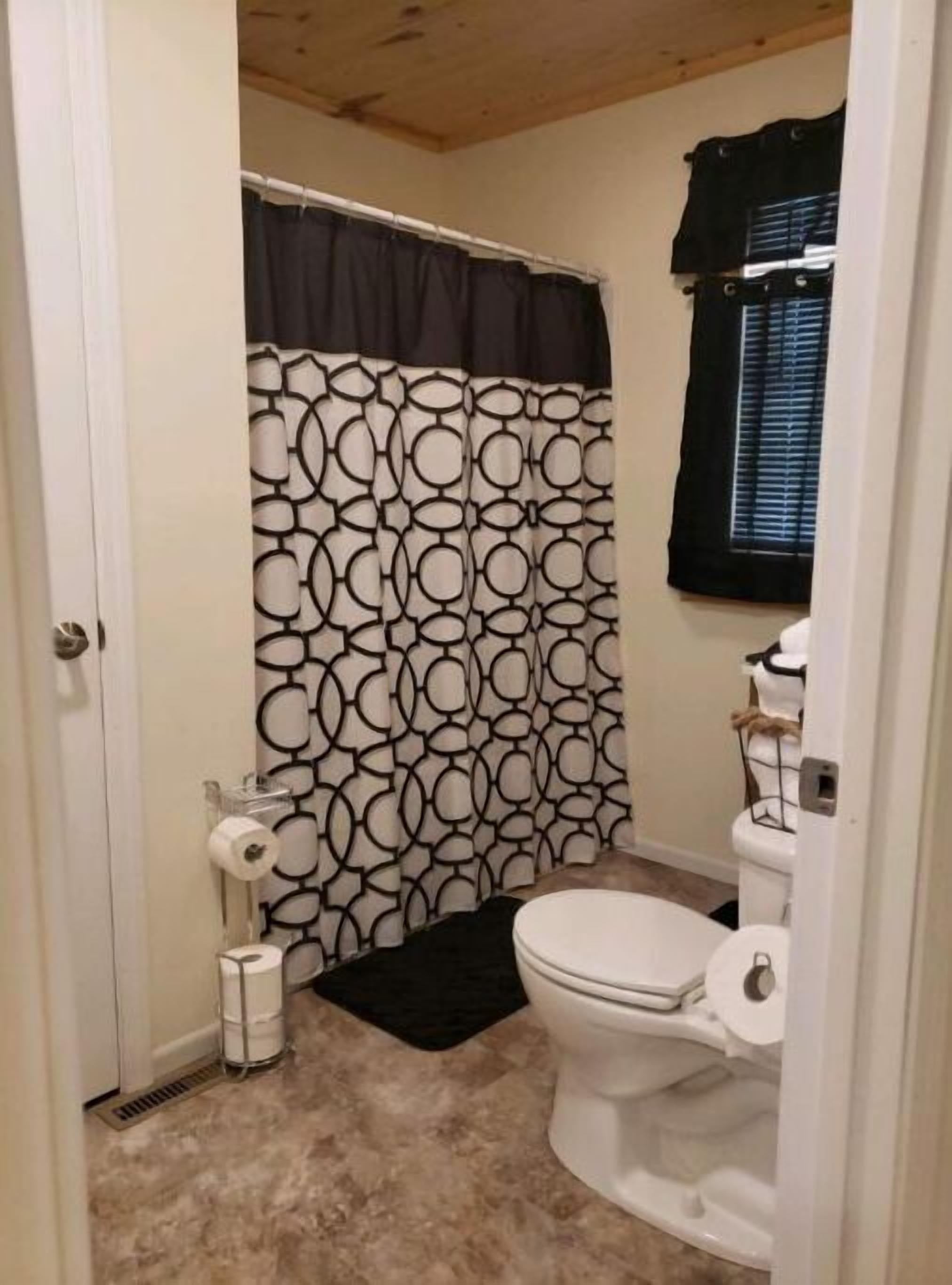 Et lille badeværelse med toilet, bruseforhæng med geometrisk mønster og et vindue med persienner.