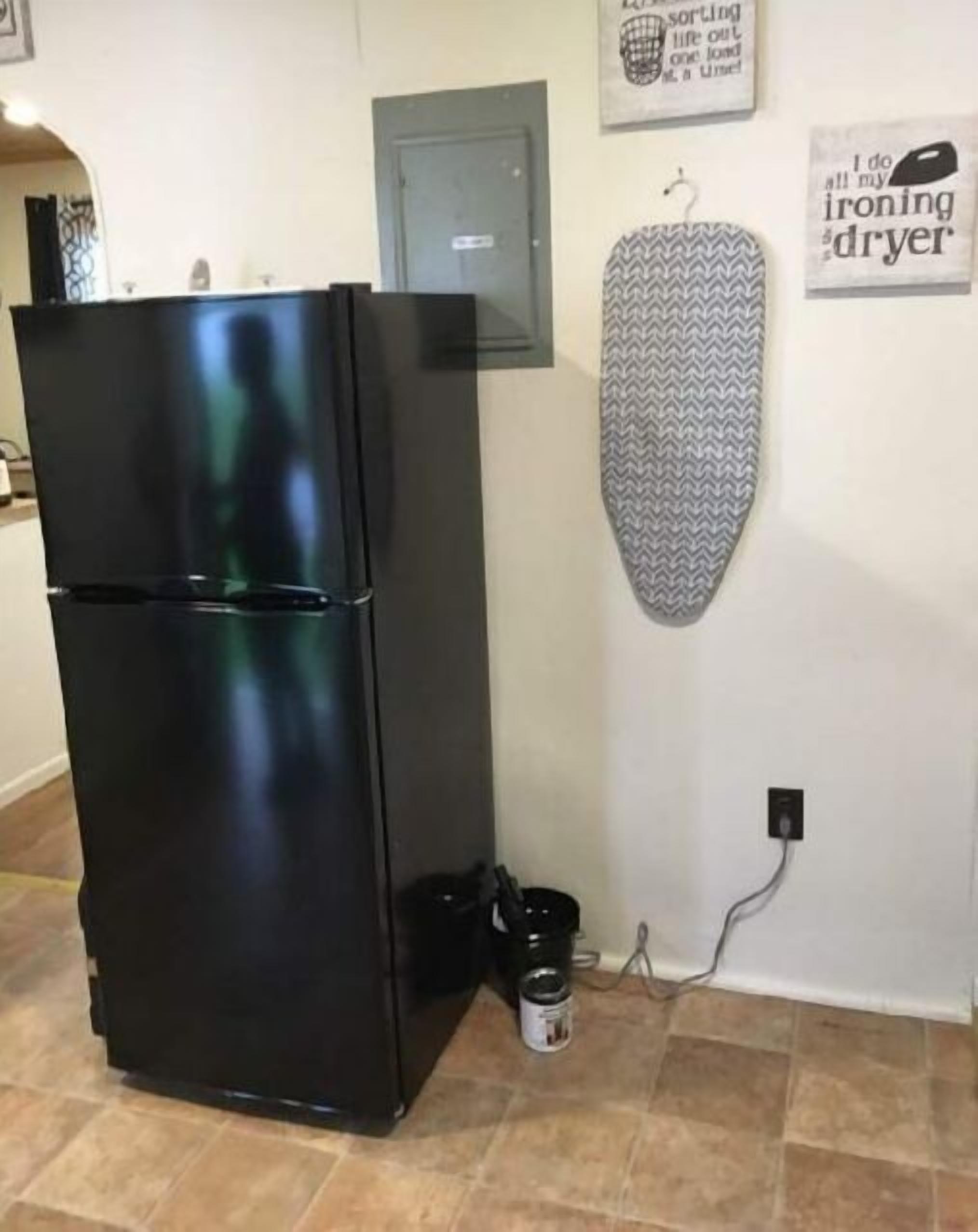 A kitchen with a black refrigerator, a grey ironing board, and a canister on the floor.