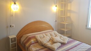 Ein ordentlich bezogenes Bett mit gestreifter Bettwäsche, ein hölzerner Kopfteil und Nachttischschränke. Zwei Wandleuchten und ein weißes Regal sind an der Wand befestigt.