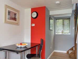 A dining area with a table, chairs, a bowl of fruit, a wall clock, and a window with blinds.